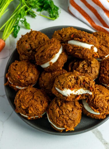 Carrot cake whoopie on a dark plate with carrots in the background.
