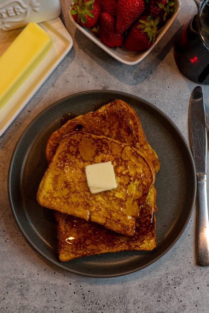 Brioche Frech toast on a dark plate with butter and maple syrup on top.