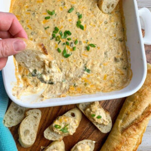 Cheesy crab dip with a some being scooped out of the white baking dish on a slice of baguette.