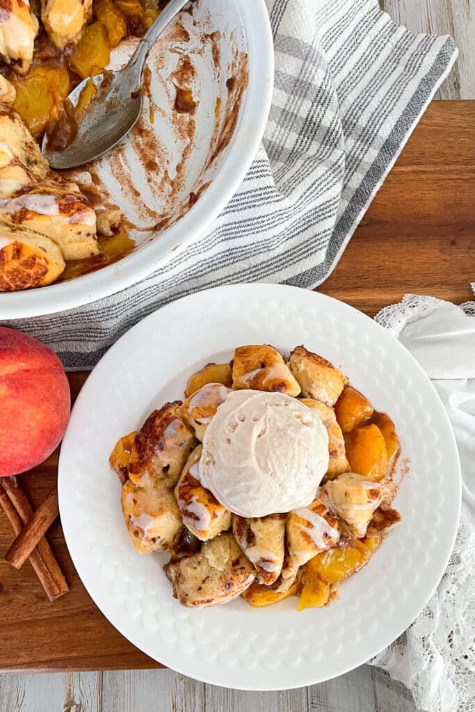 cinnamon roll peach cobbler on a serving plate with a scoop of ice cream and a peach on the side