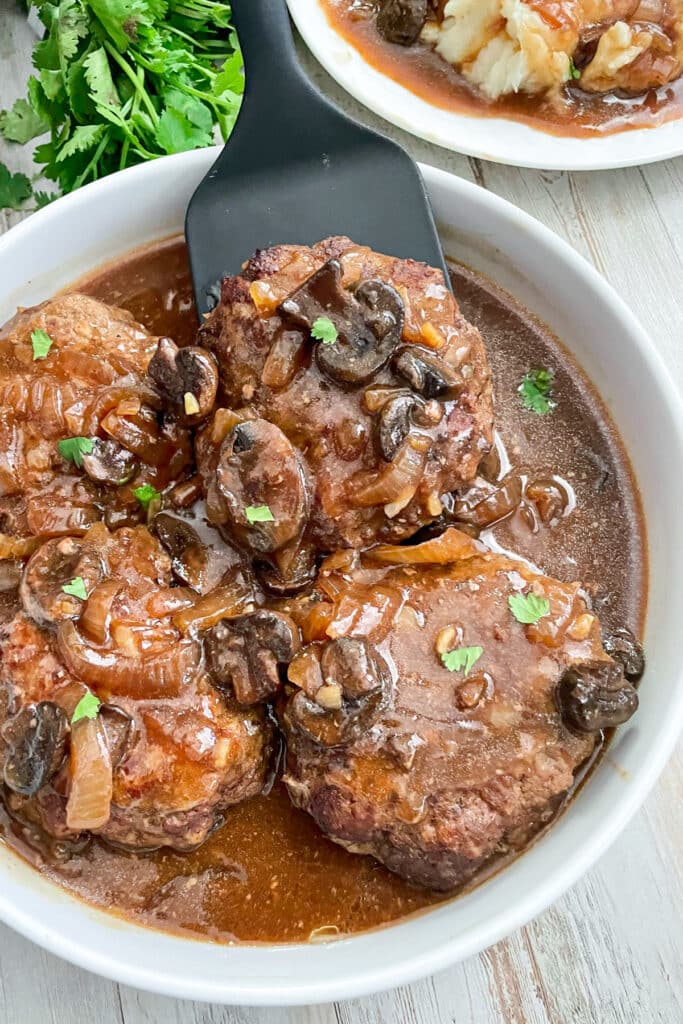 Salisbury steak patties in a white serving bowl with brown gravy, mushrooms, and onions on top