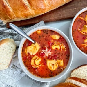 close up photo of one pot tomato tortellini soup in a large serving bowl with a spoon in the bowl. A loaf of French bread in top left hand corner with slices of French bread placed around the bowl
