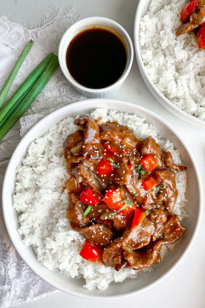 Slow cooker Beijing beef served in a white bowl with white rice. Green onions and extra sauce on the side.