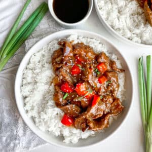 Slow cooker Beijing beef served in a white bowl with white rice. Green onions and extra sauce on the side.