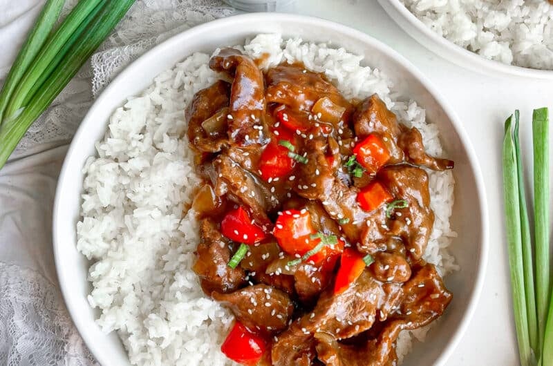 Slow cooker Beijing beef served in a white bowl with white rice. Green onions and extra sauce on the side.