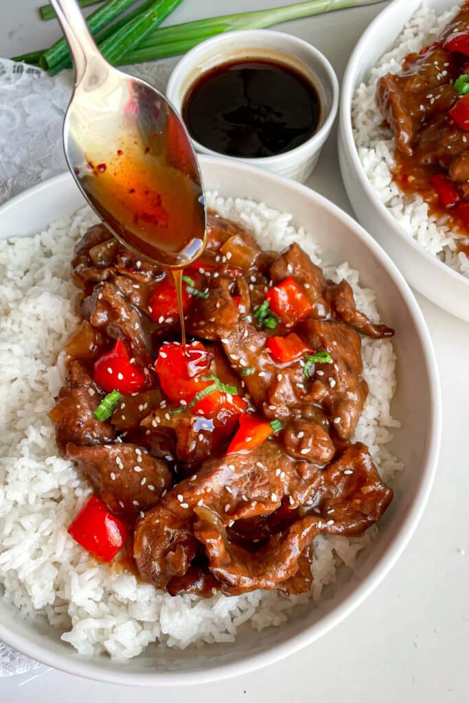 close up photo of slow cooker beijing beef in a white serving bowl with a spoon drizzeling sauce over meat