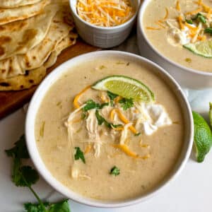 Green chicken enchilada soup in a white serving bowl topped with chedder cheese, lime wedge, and sour cream. Tortillas and shredded cheese in the background