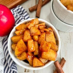 Easy Cinnamon Fried Apples served in a small white dish. A red apple and cinnamon sticks are shown next to the bowl