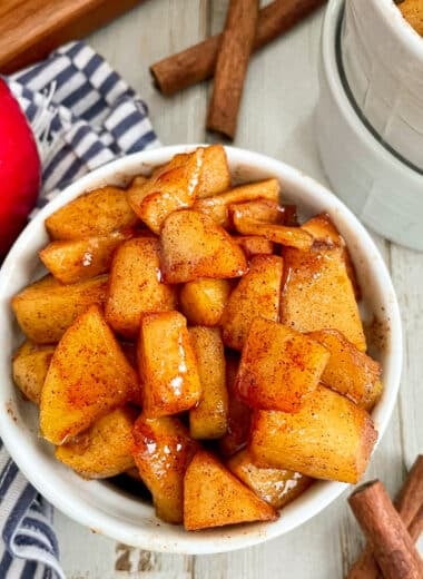 Easy Cinnamon Fried Apples served in a small white dish. A red apple and cinnamon sticks are shown next to the bowl