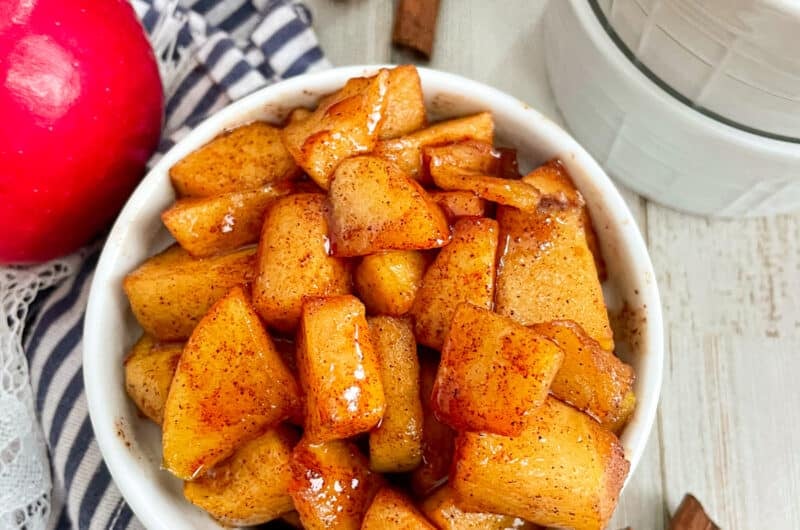 Easy Cinnamon Fried Apples served in a small white dish. A red apple and cinnamon sticks are shown next to the bowl