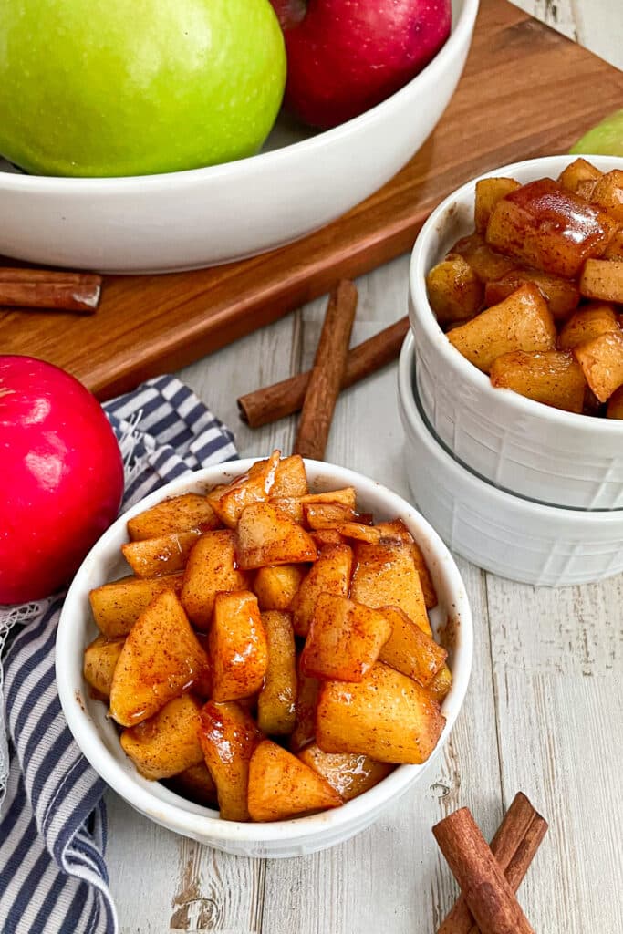 Easy Cinnamon Fried Apples served in a small white dish. A red apple and cinnamon sticks are shown next to the bowl