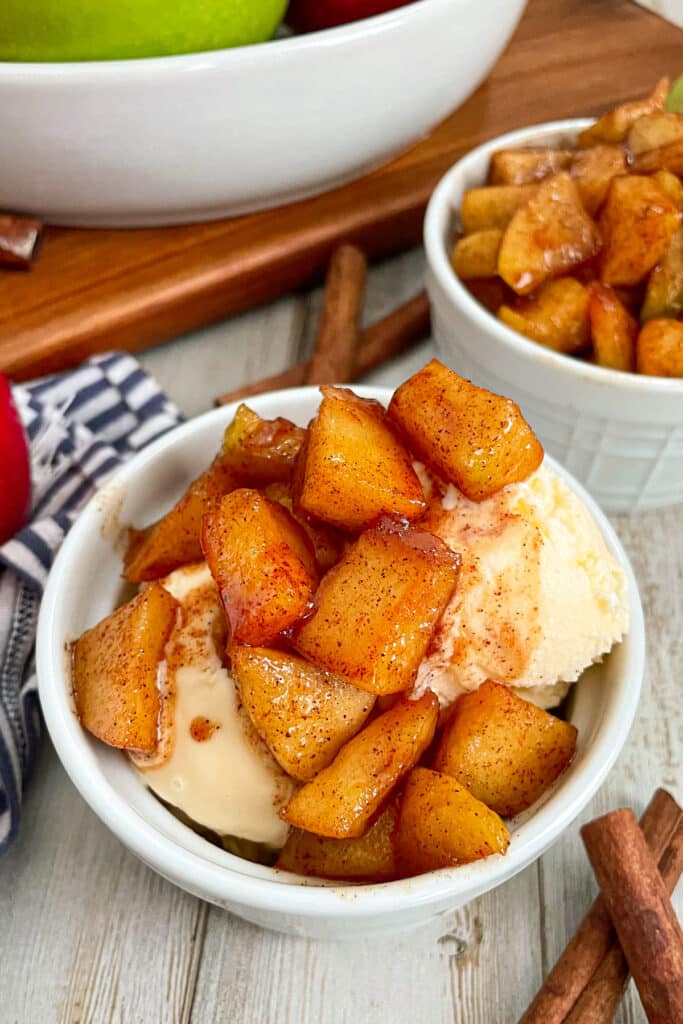Cinnamon fried apples served in a small white bowl with ice cream on the bottom and apples spooned on top. Another white bowl is shown in the back with more fried apples