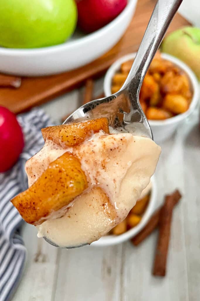 close up shot of a spoonful of vanilla ice cream and a couple bite size pieces of fried apples. A white bowl in the background with red and green apples.