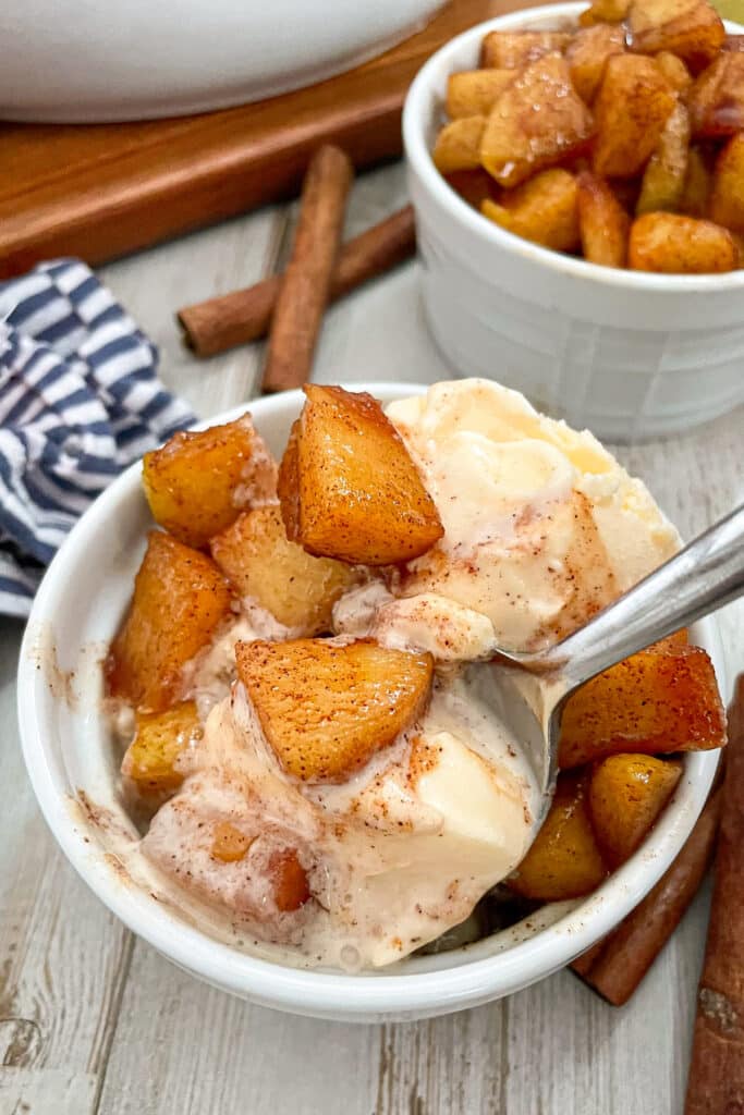 cinnamon fried apples served in a small white bowl with ice cream, apples served on top and a spoon inside. cinnamon sticks are shown next to the bowl.