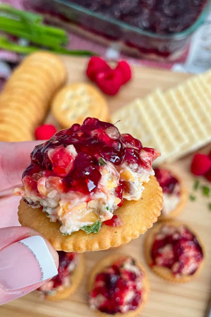 Close up shot of easy pepper jelly cheese spread on a cracker. Crackers and fresh raspberries are laid out on a cutting board