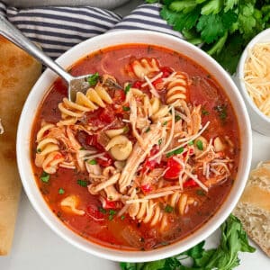 a bowl of easy chicken parmesan soup in a white serving bowl with a spoon in the left corner of the bowl. Fresh parsley lays on the side of the bowl