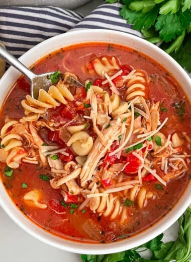 a bowl of easy chicken parmesan soup in a white serving bowl with a spoon in the left corner of the bowl. Fresh parsley lays on the side of the bowl