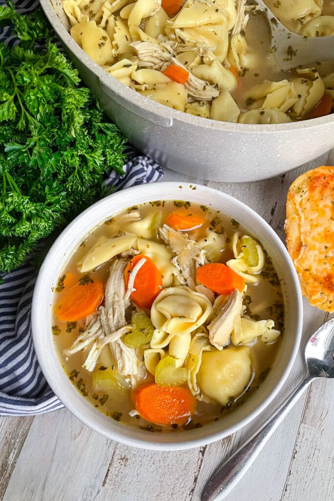 Quick & Easy chicken tortellini soup recipe in a white serving bowl. Pot of soup is behind the bowl, two pieces of bread are to the right of the bowl and parsley is to the left of the bowl.