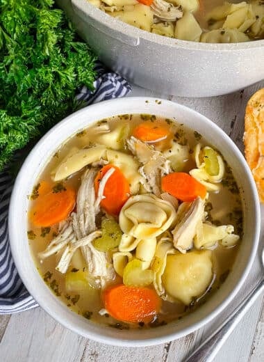 Quick & Easy chicken tortellini soup recipe in a white serving bowl. Pot of soup is behind the bowl, two pieces of bread are to the right of the bowl and parsley is to the left of the bowl.