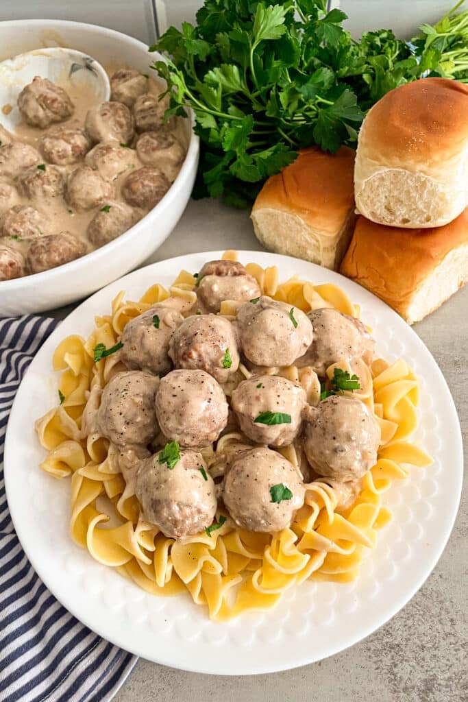 Easy Crock Pot Swedish Meatballs recipe on a white serving plate on top of cooked egg noodles for serving. Rolls and fresh parsley in the background