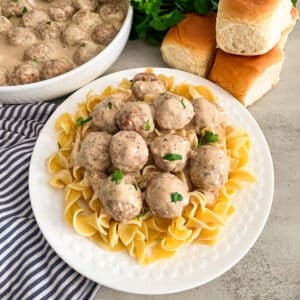 Easy Crock Pot Swedish Meatballs recipe on a white serving plate on top of cooked egg noodles for serving. Rolls and fresh parsley in the background