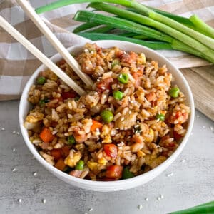 Ham fried rice in a serving bowl with chopsticks for serving. Green onions and a red bottle of hot sauce on the side