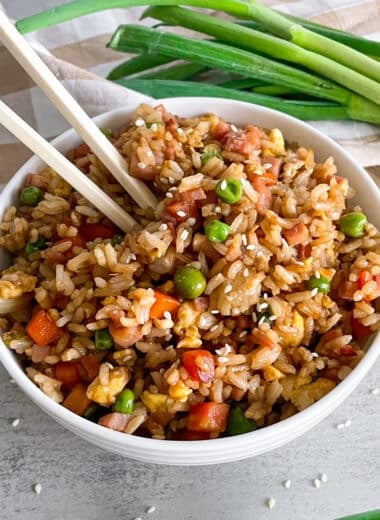 Ham fried rice in a serving bowl with chopsticks for serving. Green onions and a red bottle of hot sauce on the side