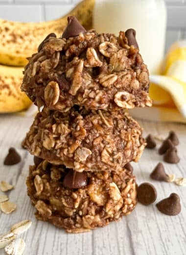 Three healthy banana oatmeal breakfast cookies stacked on top of each other. Bananas and milk shown in the background