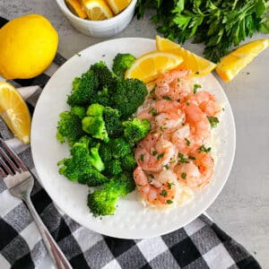 Easy oven baked shrimp on a white serving plate with steamed broccoli and sliced lemons.