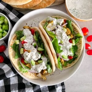 two Greek chicken gyros on a white serving plate, with tzatziki sauce sauce and pita bread in the top portion of the picture