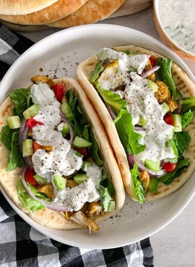 two Greek chicken gyros on a white serving plate, with tzatziki sauce sauce and pita bread in the top portion of the picture