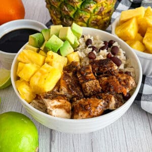 Easy Cuban Chicken and Rice Bowl (with Black Beans) in a white serving bowl. Fresh pineapple, oranges, limes, and cuban sauce place above the bowl in the pictures.
