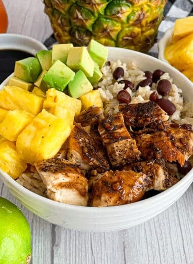 Easy Cuban Chicken and Rice Bowl (with Black Beans) in a white serving bowl. Fresh pineapple, oranges, limes, and cuban sauce place above the bowl in the pictures.