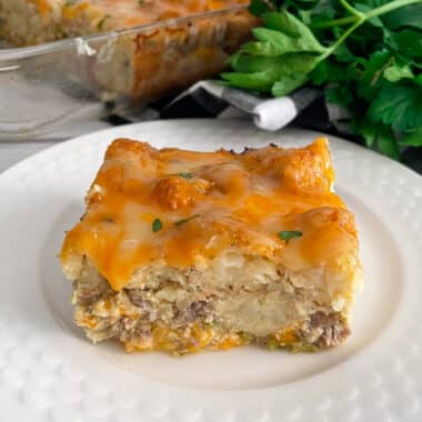 slice of tator tot breakfast casserole on a white serving plate with fresh parsley and casserole dish in the background