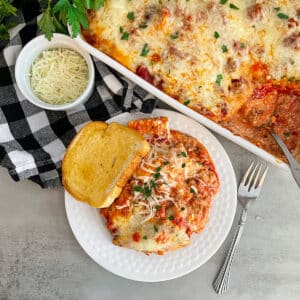 Baked cheese ravioli casserole on a white serving plate with garlic toast and fresh parmesan cheese on the side.