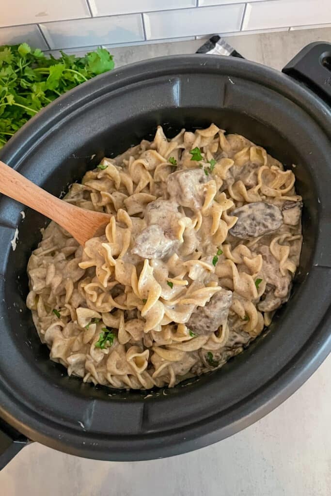 beef stroganoff in a black slow cooker. A wooden spoon is for serving and fresh basil sprinkled on top.