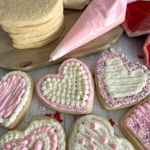 Valentines day cookies frosted and topped with sprinkles. Piping bag with pink frosting and unfrosted cookies in the background