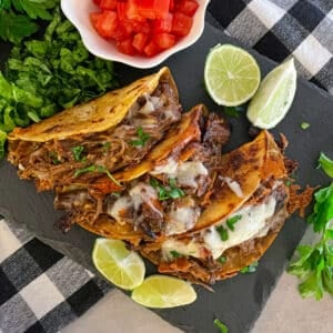 Easy crock pot shredded beef tacos on a black serving plate with a side of chopped tomatoes, sliced limes, and cilantro