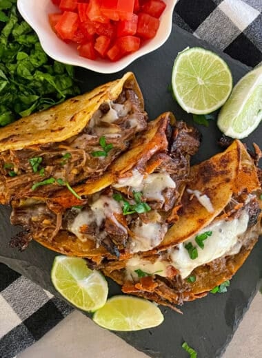 Easy crock pot shredded beef tacos on a black serving plate with a side of chopped tomatoes, sliced limes, and cilantro