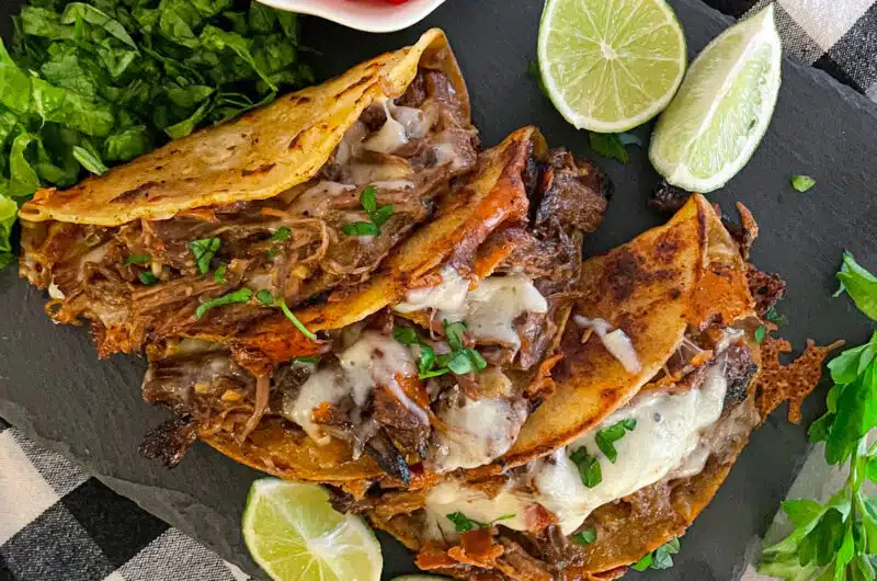 Easy crock pot shredded beef tacos on a black serving plate with a side of chopped tomatoes, sliced limes, and cilantro