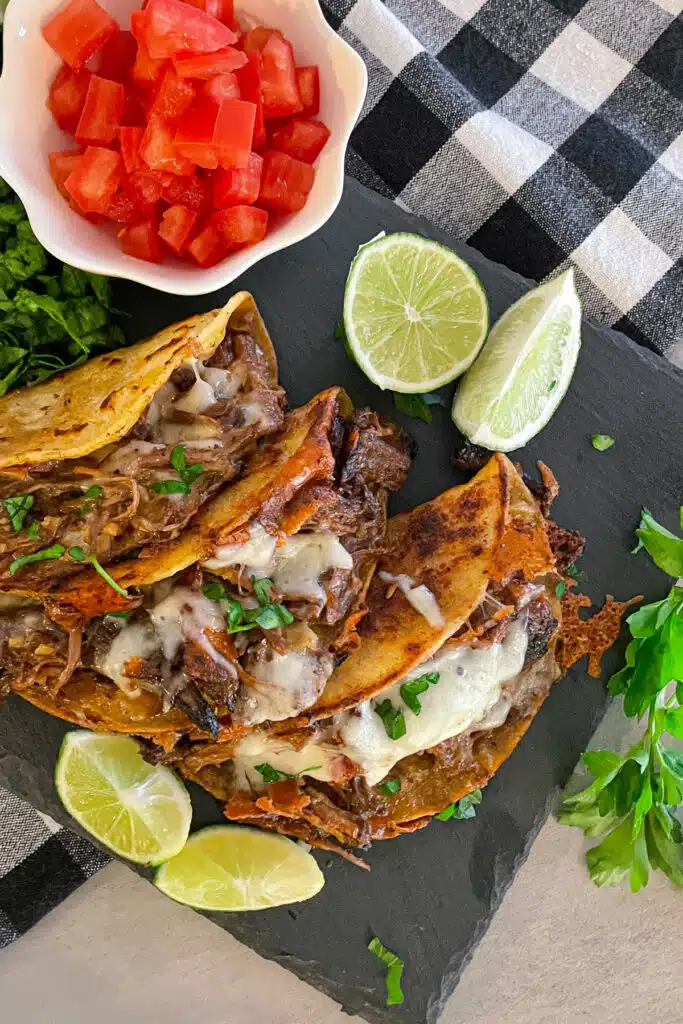 Easy crock pot shredded beef tacos on a black serving plate with a side of chopped tomatoes, sliced limes, and cilantro