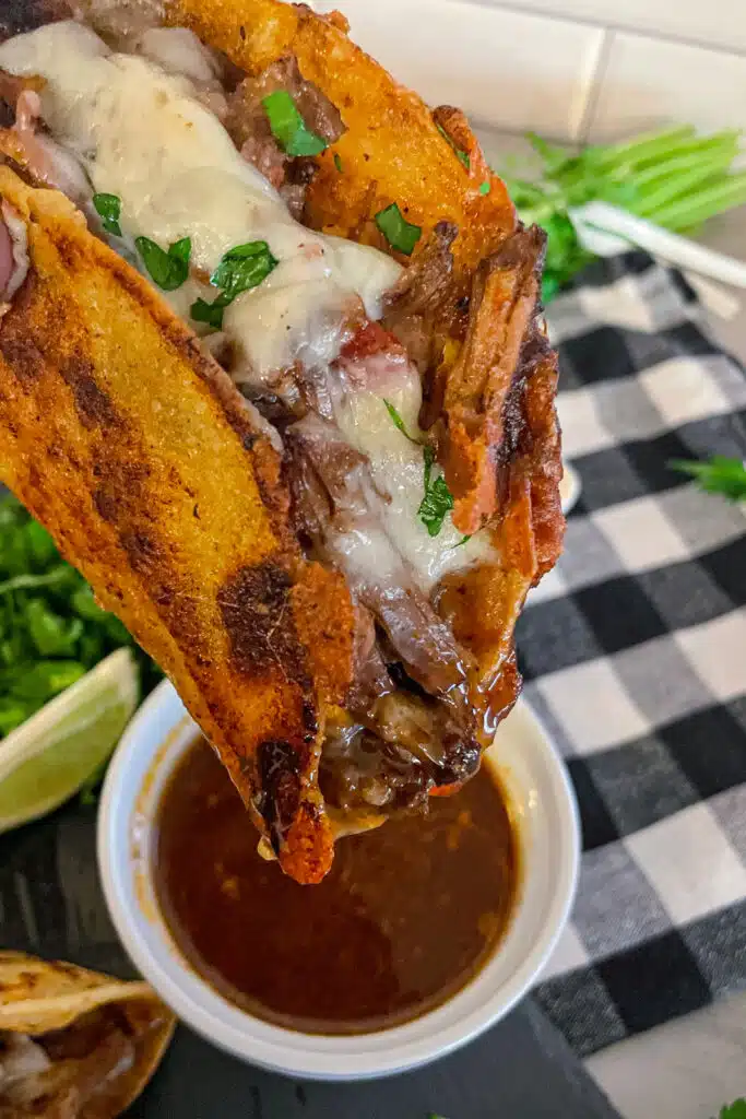 close up photo of a shredded beef taco with a crispy corn tortilla topped with shredded cheese being dipped in consume for eating