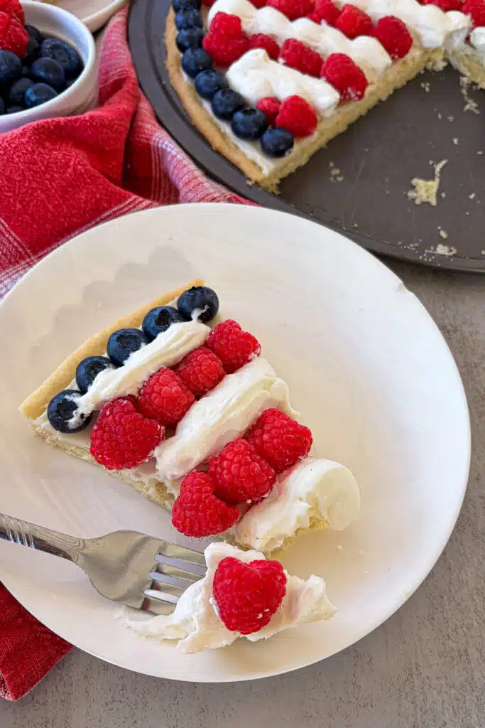 one slice of fruit pizza placed on a white plate for eating. A single fork took a bite out of it. Berries in the background.